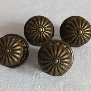 Może przedstawiać: Cztery antyczne mosiężne gałki do szafek. Każda gałka ma okrągły, kwiatowy wzór z centralnym okrągłym detalem. Gałki mają ciepłą, postarzaną patynę, co sugeruje, że są w stylu vintage. Ułożone są na białej powierzchni.