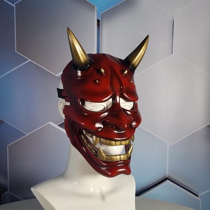May include: A red and gold Oni mask with horns, sharp teeth, and a fierce expression. The mask is on a white mannequin head.