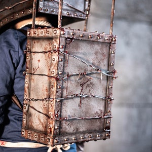 May include: A person wearing a costume with a rusty metal box on their back. The box is covered in barbed wire and has blood splatters on it.