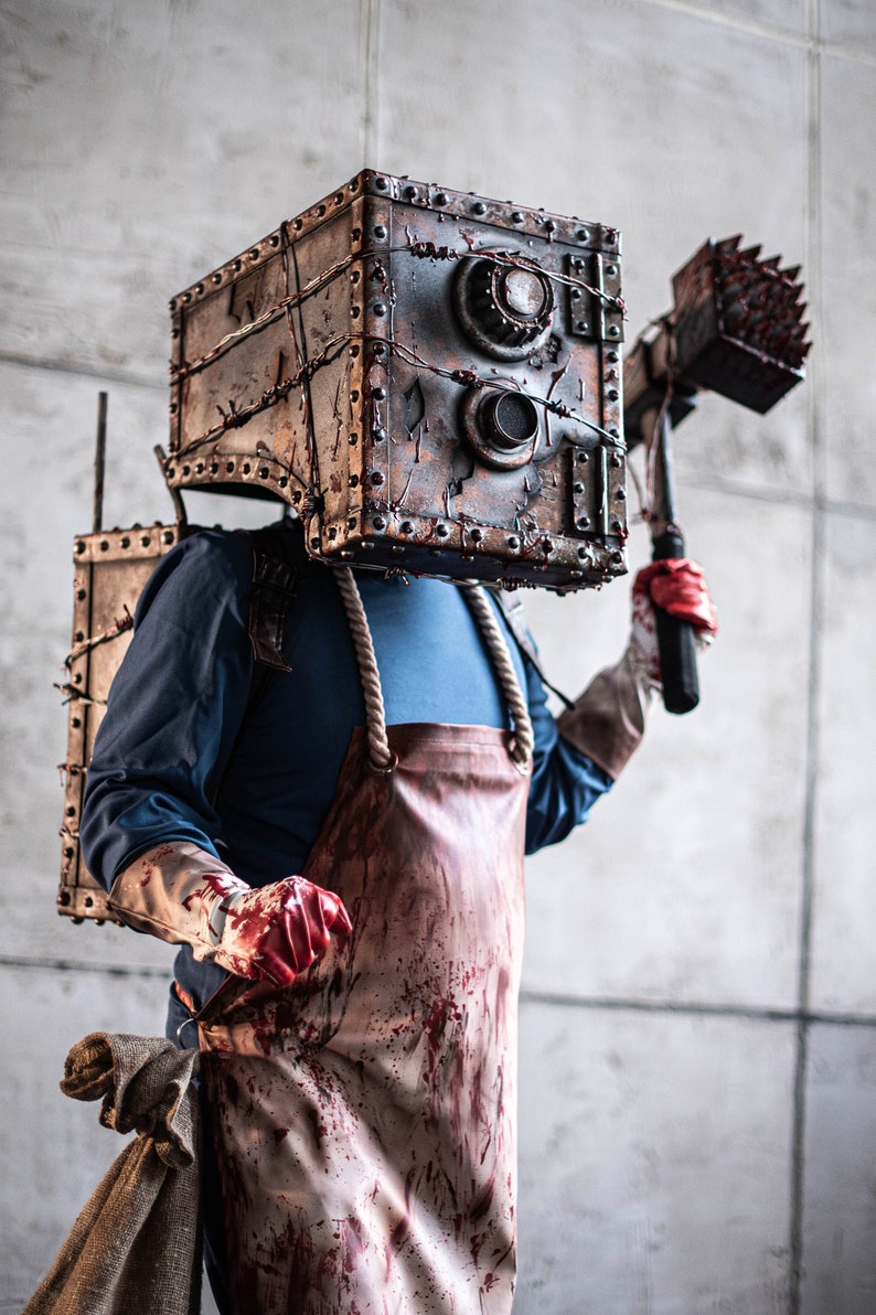 May include: A person wearing a blood-stained apron and a metal box on their head, holding a spiked hammer. The box has two circular openings and is covered in barbed wire.