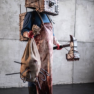May include: A person wearing a bloody butcher's apron and a metal box on their head, holding a large spiked axe and a burlap sack with hooks sticking out.
