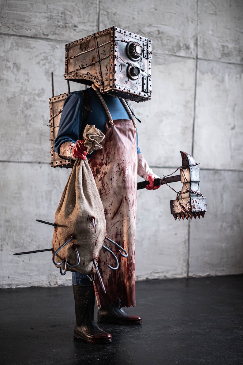 May include: A person wearing a bloody butcher's apron and a metal box on their head, holding a large spiked axe and a burlap sack with hooks sticking out.