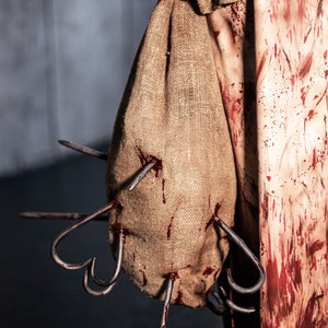 May include: A burlap sack with blood stains and multiple metal hooks protruding from it. The sack is being held by a person wearing a white and red stained apron.