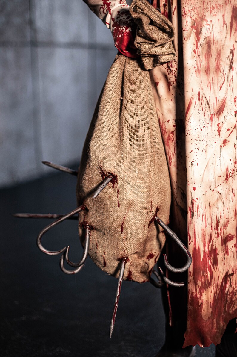 May include: A burlap sack with blood stains and multiple metal hooks protruding from it. The sack is being held by a person wearing a white and red stained apron.
