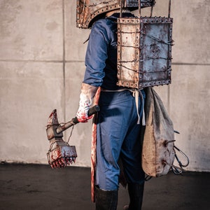 May include: A person wearing a metal box over their head and a metal chest plate with barbed wire. They are holding a bloody hammer with a spiked head. The person is wearing blue overalls and black boots.
