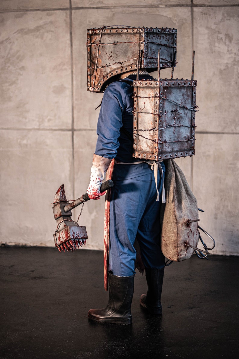 May include: A person wearing a metal box over their head and a metal chest plate with barbed wire. They are holding a bloody hammer with a spiked head. The person is wearing blue overalls and black boots.