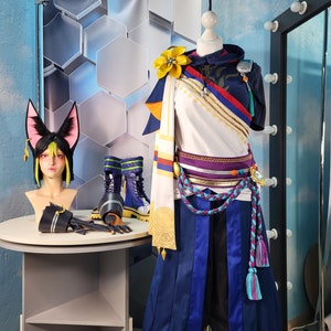 Può includere: Un costume blu e bianco con un fiore giallo, una testa di gatto bianco e nero e stivali neri e blu. Il costume ha una cintura con una fibbia dorata e una lunga sciarpa blu e bianca.