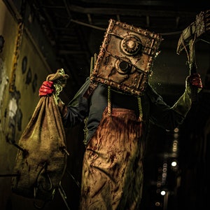 May include: A person wearing a bloody apron and a metal box mask holds a burlap sack and a rusty axe. The person is standing in a dark hallway with peeling paint on the walls.