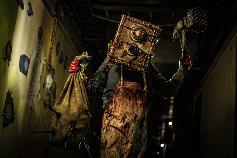 May include: A person wearing a bloody apron and a metal box mask holds a burlap sack and a rusty axe. The person is standing in a dark hallway with peeling paint on the walls.