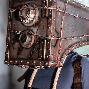 May include: A person wearing a rusty metal mask with barbed wire and blood splatters. The mask is shaped like a safe with two round dials. The person is wearing a blue shirt and a brown leather strap.