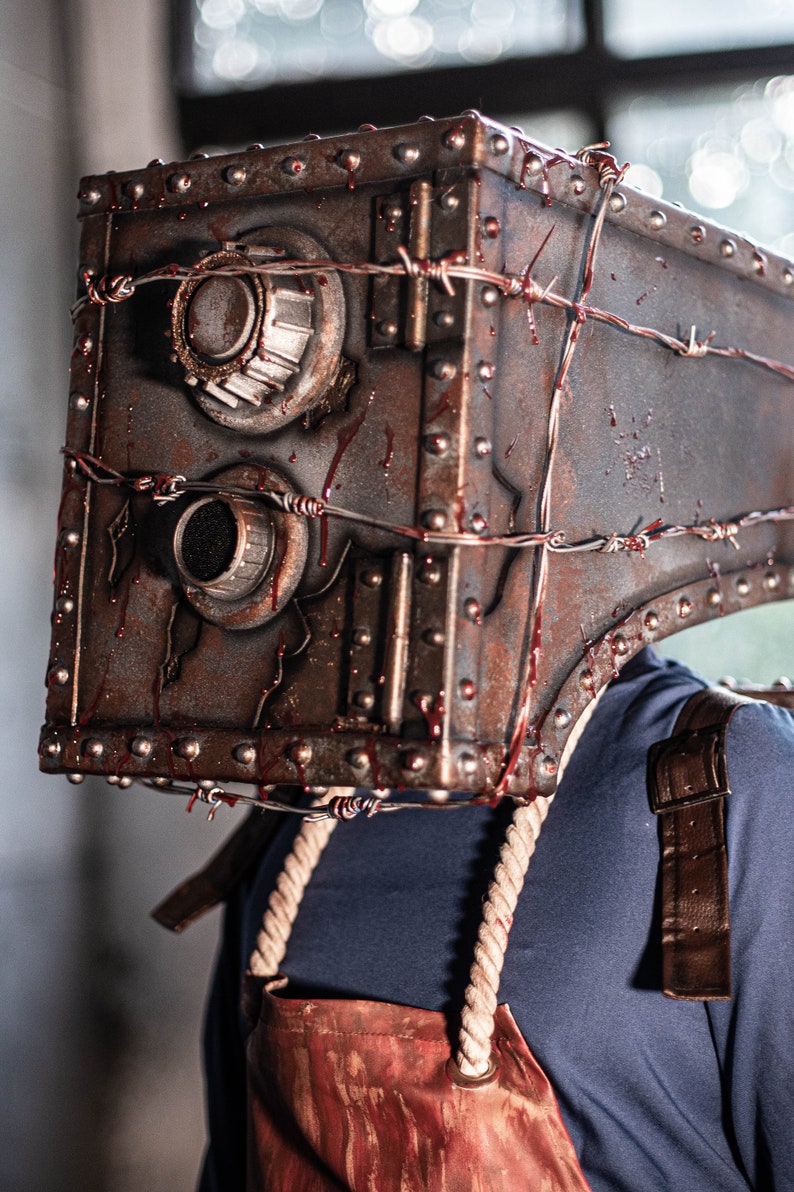 May include: A person wearing a rusty metal mask with barbed wire and blood splatters. The mask is shaped like a safe with two round dials. The person is wearing a blue shirt and a brown leather strap.