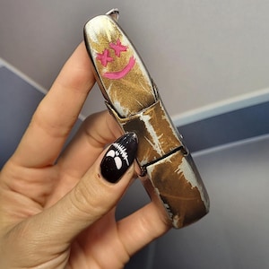 May include: A gold-colored lighter with a pink smiley face drawn on it. The lighter is held in a hand with black nail polish and white skull designs.