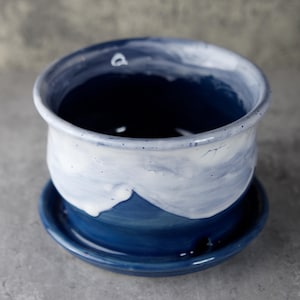 May include: A handcrafted ceramic planter with a matching saucer. The planter is a deep blue color with a white and blue wave-like design around the rim. The saucer is a solid, dark blue. This decorative piece is ideal for small plants.