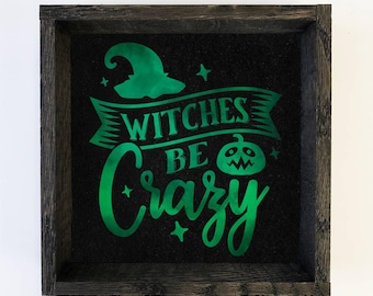 May include: A square wooden sign with a dark frame and a black background. The sign features the phrase "Witches Be Crazy" in bright green lettering, along with a witch hat and a jack-o'-lantern. The sign is a Halloween decoration.