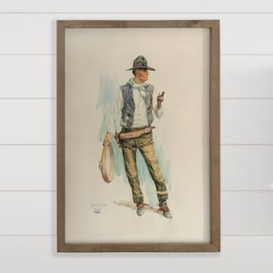 May include: Watercolor print of a cowboy in a hat, vest, and plaid pants, holding a lasso. The artwork is framed in a wooden frame. The cowboy is standing in a relaxed pose, with a light blue background.