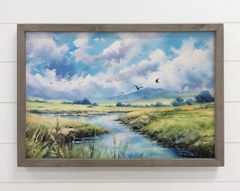 May include: A framed landscape painting depicts a serene river scene under a cloudy blue sky. The artwork features green fields, distant mountains, and birds in flight, all within a gray wooden frame.