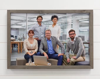 Könnte beinhalten: Gerahmtes Foto einer Gruppe von fünf Personen in einem modernen Büro. Sie tragen Business-Casual-Kleidung, mit einem Laptop im Vordergrund. Der Rahmen ist grau und das Bild hängt an einer weißen Wand.