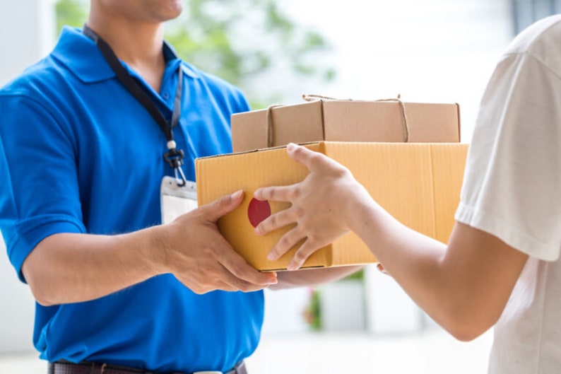 May include: A delivery person in a blue polo shirt hands two brown cardboard boxes to a person wearing a white shirt.