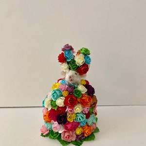May include: A small white mouse wearing a crown of colorful paper flowers. The mouse is surrounded by a large bouquet of colorful paper flowers.