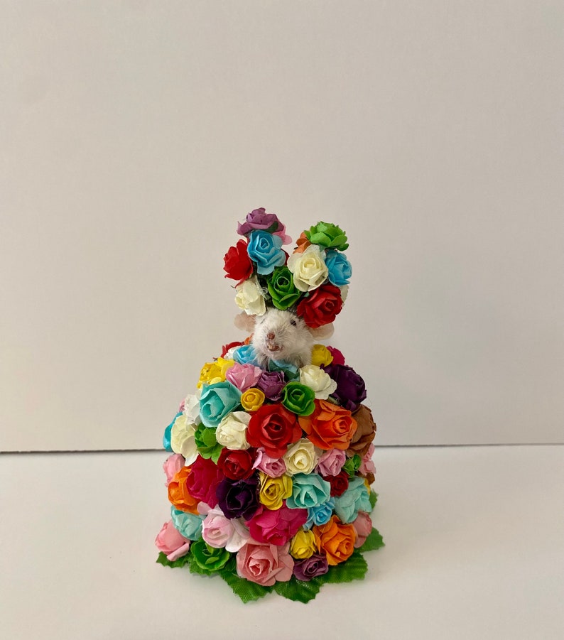 May include: A small white mouse wearing a crown of colorful paper flowers. The mouse is surrounded by a large bouquet of colorful paper flowers.
