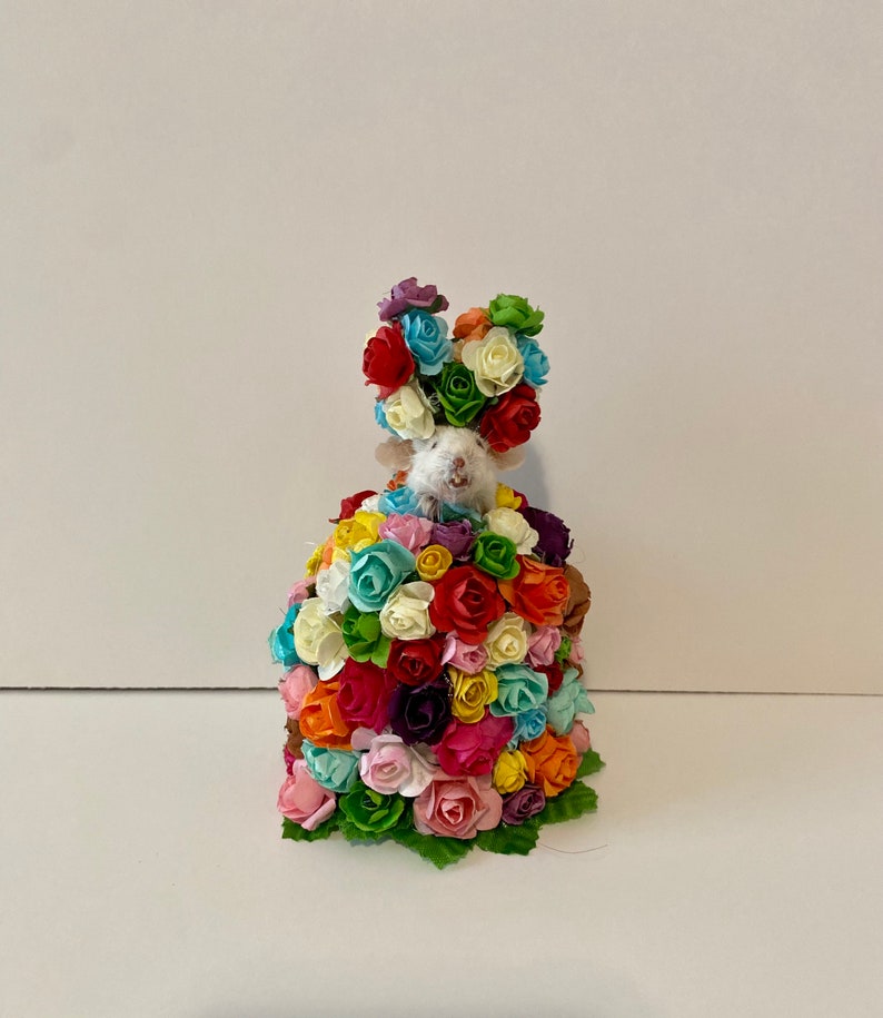 May include: A white mouse figurine wearing a crown of colorful paper flowers. The mouse is surrounded by a large bouquet of colorful paper flowers.