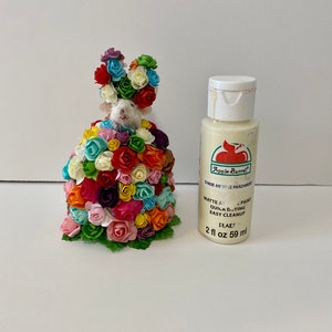 May include: A small white mouse figurine covered in colorful paper flowers. The mouse is wearing a crown of flowers. A bottle of white paint labeled "Apple Barrel" is next to the mouse.