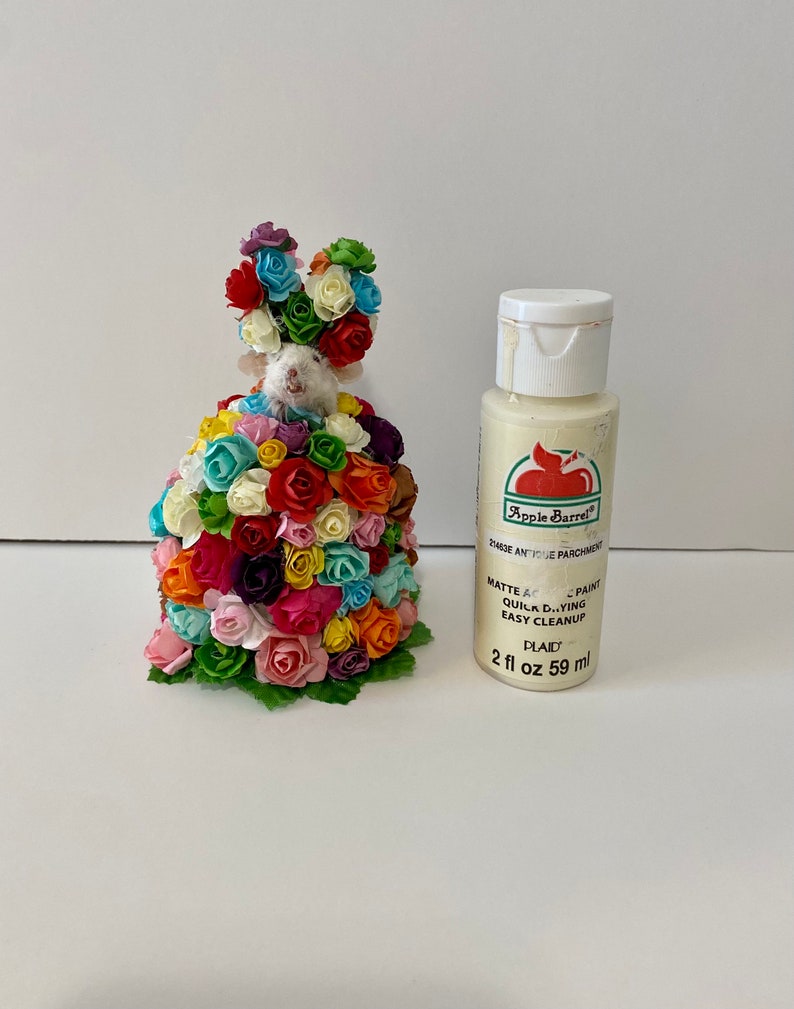 May include: A small white mouse figurine covered in colorful paper flowers. The mouse is wearing a crown of flowers. A bottle of white paint labeled "Apple Barrel" is next to the mouse.