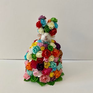 May include: A white mouse wearing a crown of colorful artificial flowers. The mouse is surrounded by a large bouquet of colorful artificial roses.