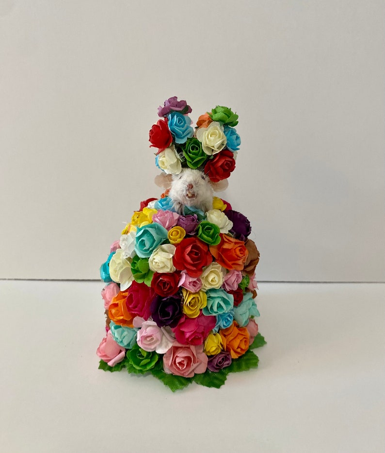 May include: A white mouse wearing a crown of colorful artificial flowers. The mouse is surrounded by a large bouquet of colorful artificial roses.