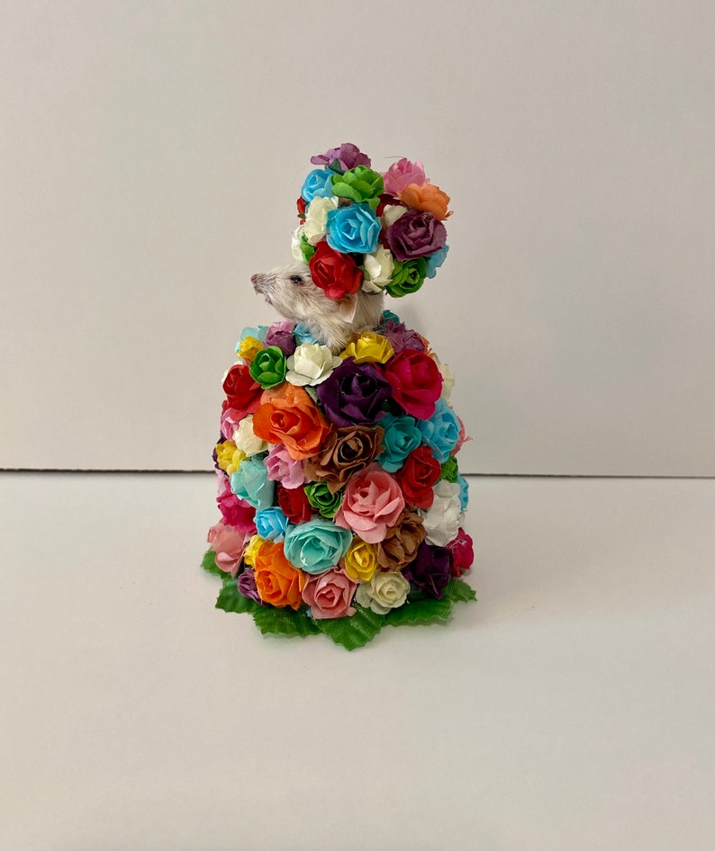 May include: A small white mouse wearing a crown of colorful paper flowers. The mouse is surrounded by a large bouquet of colorful paper flowers.