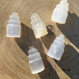 May include: Five white selenite crystal towers, each with a layered, stepped design, arranged on a light brown wooden surface.
