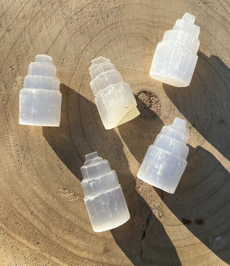 May include: Five white selenite crystal towers, each with a layered, stepped design, arranged on a light brown wooden surface.