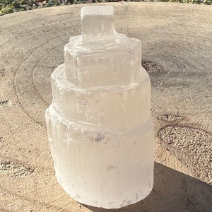 May include: A white selenite crystal tower with a layered, tiered design. The crystal is smooth and translucent, with a slightly cloudy appearance.