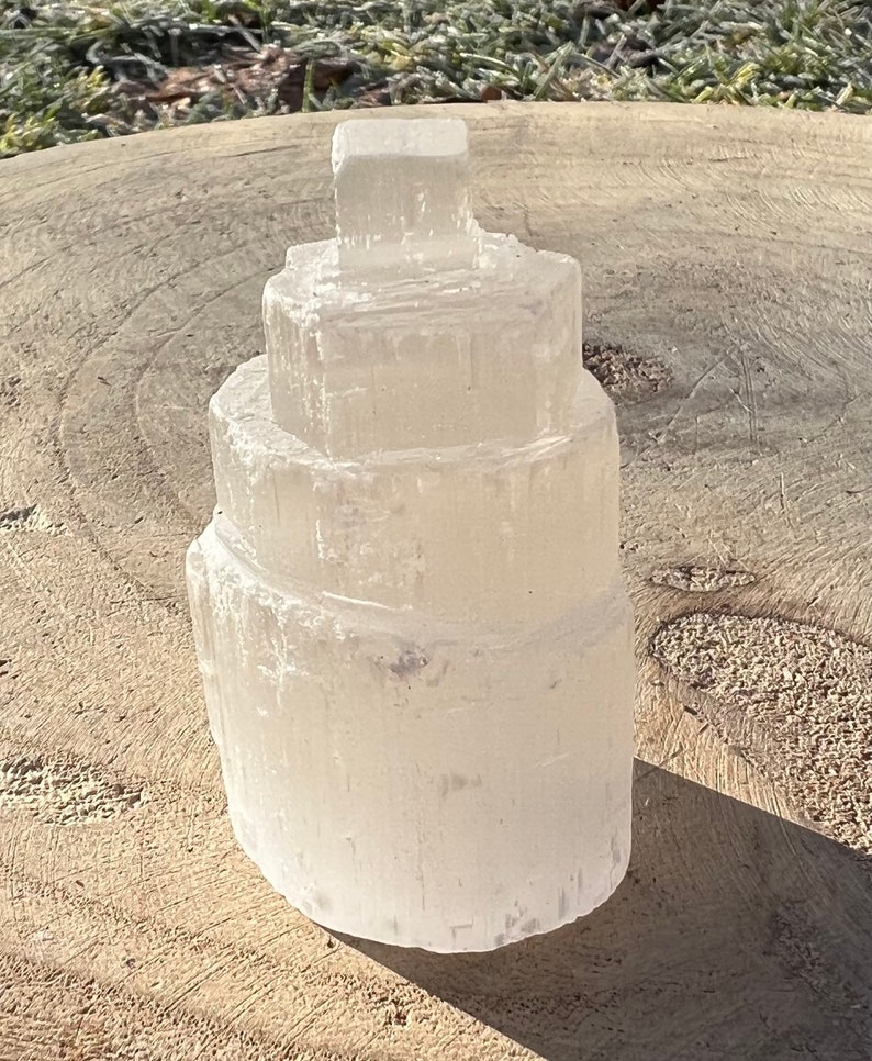 May include: A white selenite crystal tower with a layered, tiered design. The crystal is smooth and translucent, with a slightly cloudy appearance.