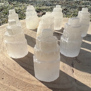 May include: A collection of seven white selenite crystal towers of varying sizes, arranged on a light brown wooden surface. The crystals are stacked in a tiered formation, resembling small towers.