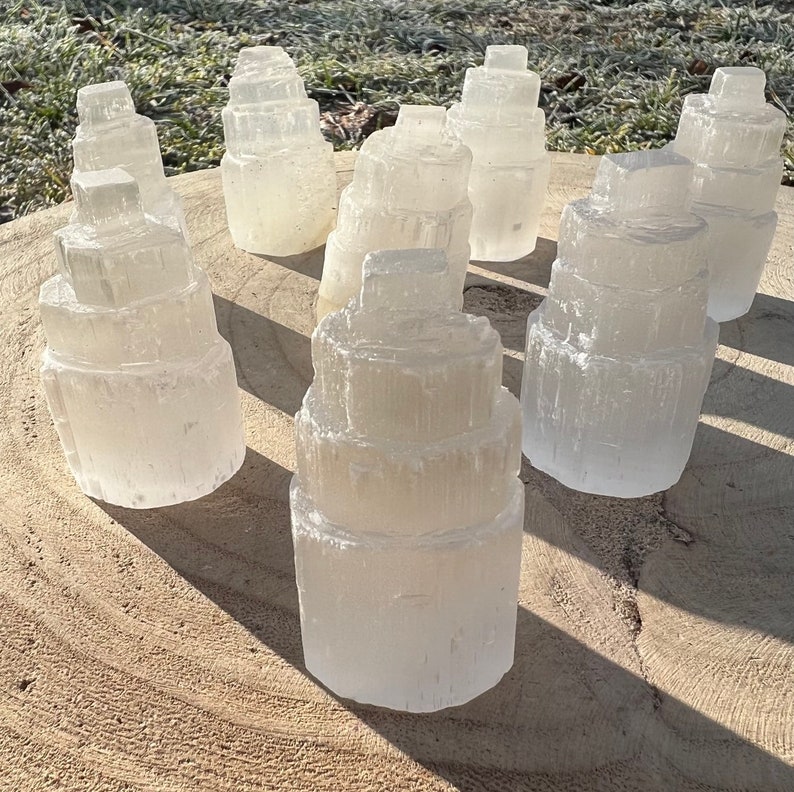 May include: A collection of seven white selenite crystal towers of varying sizes, arranged on a light brown wooden surface. The crystals are stacked in a tiered formation, resembling small towers.
