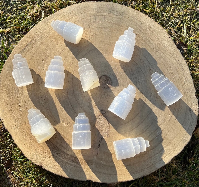 May include: A collection of ten selenite crystal towers, each with a unique shape and size, arranged on a wooden surface. The crystals are white and translucent, with a smooth, polished finish.