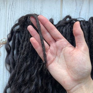 May include: A close-up of a hand holding a thick strand of black dreadlocks. The dreadlocks are long and curly, and the hand is holding them up to show their texture.