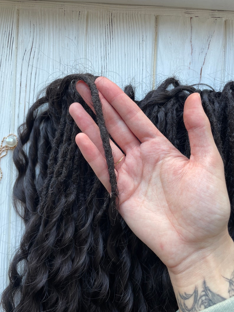 May include: A close-up of a hand holding a thick strand of black dreadlocks. The dreadlocks are long and curly, and the hand is holding them up to show their texture.
