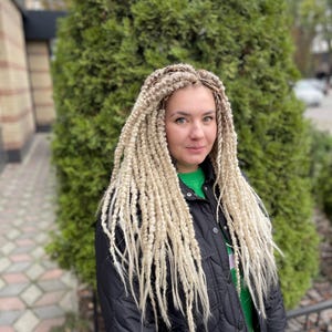 May include: A woman with long, blonde braids wearing a black jacket over a green shirt.