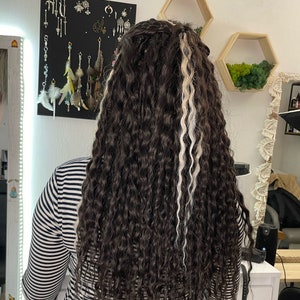 May include: A person with long, curly brown hair with two white streaks. The hair is styled in a ponytail. The person is wearing a black and white striped shirt.