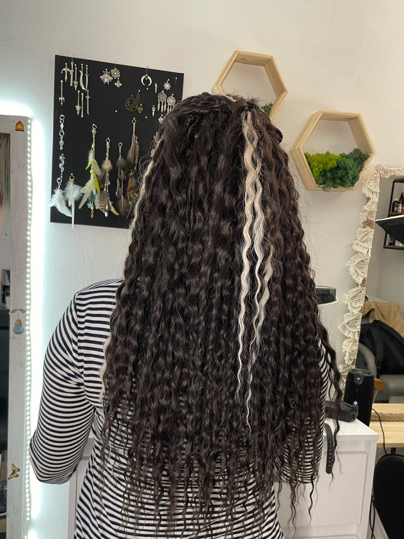 May include: A person with long, curly brown hair with two white streaks. The hair is styled in a ponytail. The person is wearing a black and white striped shirt.