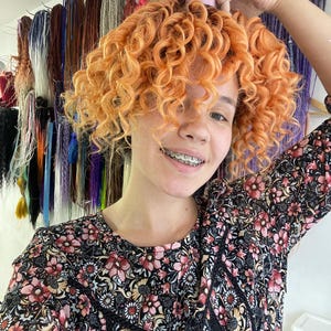 May include: A person with orange curly hair wearing a black floral dress and braces. The person is holding a section of their hair up with a pink hair tie.