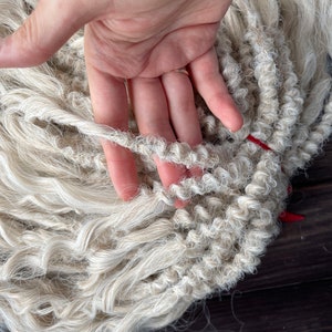 May include: A close-up of a bundle of white, curly, synthetic hair extensions. The hair is arranged in a loose braid and is soft and fluffy.