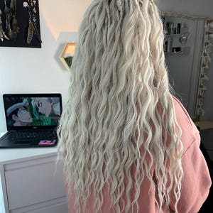 May include: A person with long, wavy, platinum blonde hair with small, white beads woven into the hair. The person is wearing a light pink sweatshirt.