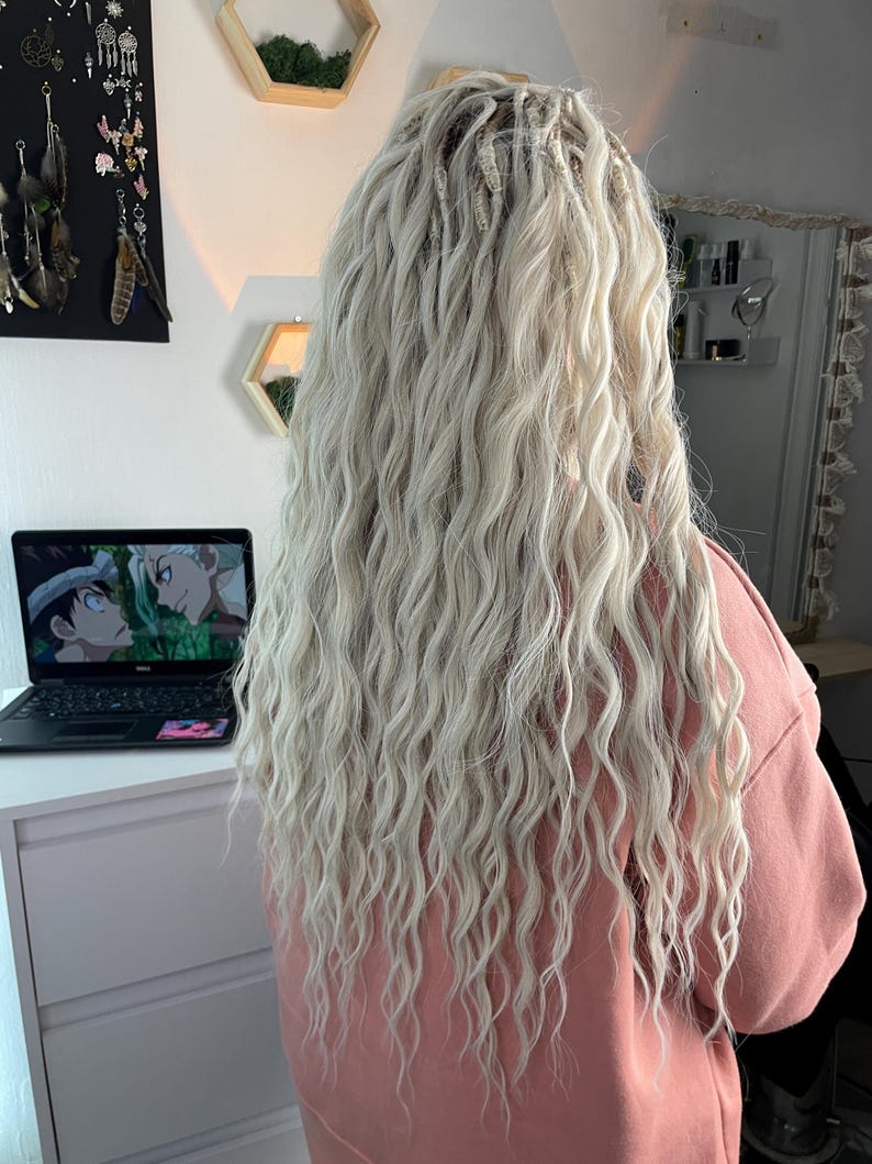 May include: A person with long, wavy, platinum blonde hair with small, white beads woven into the hair. The person is wearing a light pink sweatshirt.
