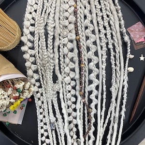 Puede incluir: Una colección de rastas sintéticas blancas con diferentes texturas y adornos, incluyendo cuentas y dijes. Las rastas se muestran en una bandeja negra.