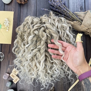 May include: A large bundle of curly, light blonde and white human hair extensions. The hair is arranged in a loose, messy bun.