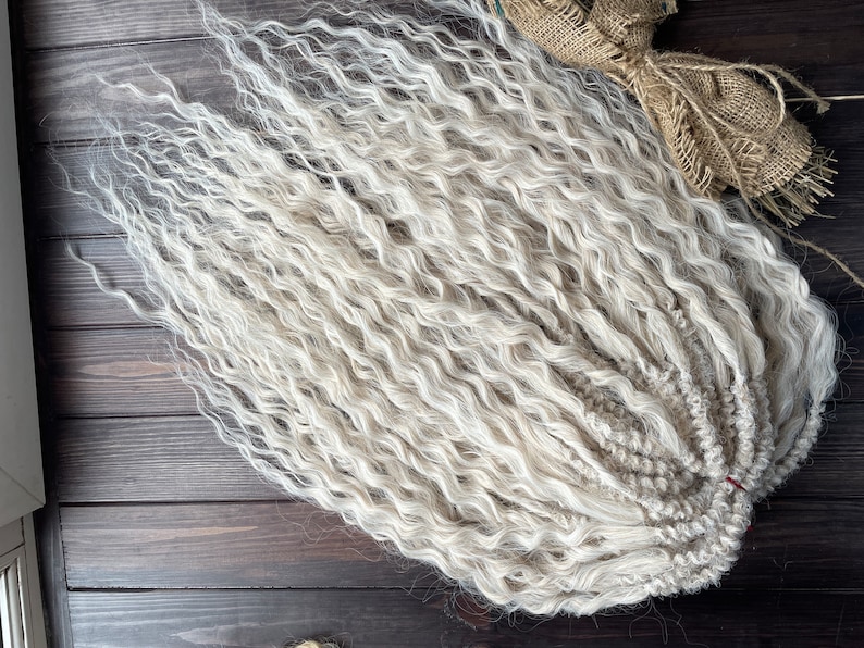 May include: A bundle of long, white, wavy synthetic hair extensions. The hair is arranged in a loose braid.