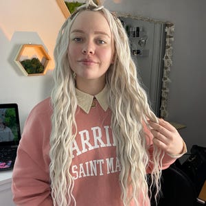 May include: A woman with long, wavy, blonde hair is wearing a pink sweatshirt with the text "Warriors Saint Mary" printed on the front.
